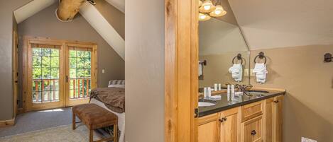 A bedroom with a large window, a wooden bench, and a bathroom with a sink and mirror.