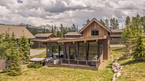 A modern wooden cabin with a deck, surrounded by trees and mountains.
