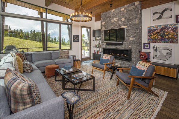 A modern living room with a stone fireplace, large windows, and a view of a green landscape. - Luxury Moonlight Basin Cabin with Ski-In/Ski-Out Access, Hot Tub & Mountain Views (Big Sky)