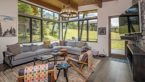 A modern living room with a sectional sofa, a coffee table, and a fireplace. Large windows offer a view of the outdoors.