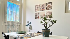 Un comedor con una mesa blanca, sillas negras, una planta verde en una maceta trenzada y un mantel decorado.