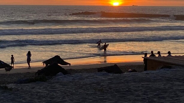 Escena de playa con el sol poniéndose sobre el océano, personas caminando por la orilla y un rompeolas rocoso en primer plano.