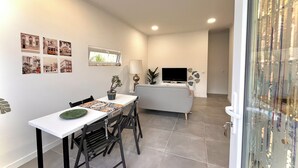 Una sala moderna con una televisión de pantalla plana, un comedor con una mesa blanca y sillas negras, y un pequeño balcón con una planta en maceta.