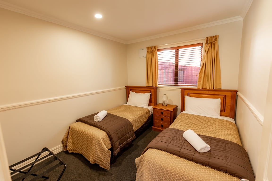 A hotel room with two beds, a window with curtains, a bedside table with a lamp, and a radiator.