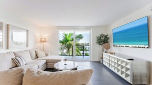 Uma sala de estar moderna com sofá, mesa de centro e uma grande televisão exibindo uma cena de praia.
