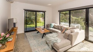 A modern living room with a sofa, coffee table, and a TV on a wooden stand. There are sliding glass doors leading to a balcony with a view of greenery. - 1 Anderson Ave Sandy Point (Sandy Point)
