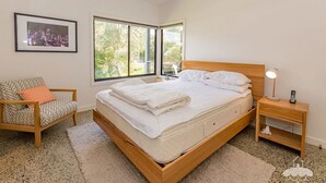 A neatly made bed with white linens, a wooden nightstand with a lamp and phone, a chair with patterned cushions, and a large window offering a view of greenery. - 1 Anderson Ave Sandy Point (Sandy Point)