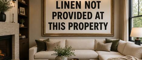 Ein Wohnzimmer mit einer Couch, einem Couchtisch mit Büchern und einer Kerze, einem Kamin und einem gerahmten Schild mit der Aufschrift 'LINEN NOT PROVIDED AT THIS PROPERTY'.