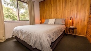 A bedroom with a large bed, wooden panel walls, a window with curtains, and a bedside table with a lamp.