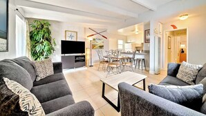 A modern living room with a sofa, a coffee table, and a TV. There is a dining area with a table and chairs. A large plant is in the corner.