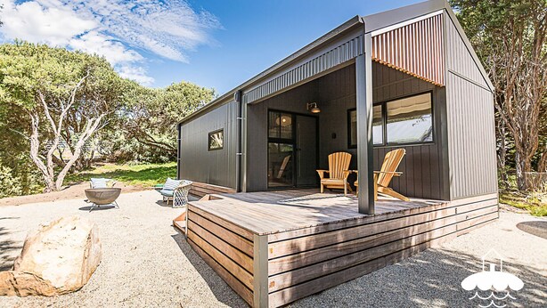 A modern cabin with a wooden deck, outdoor seating, and a large window.