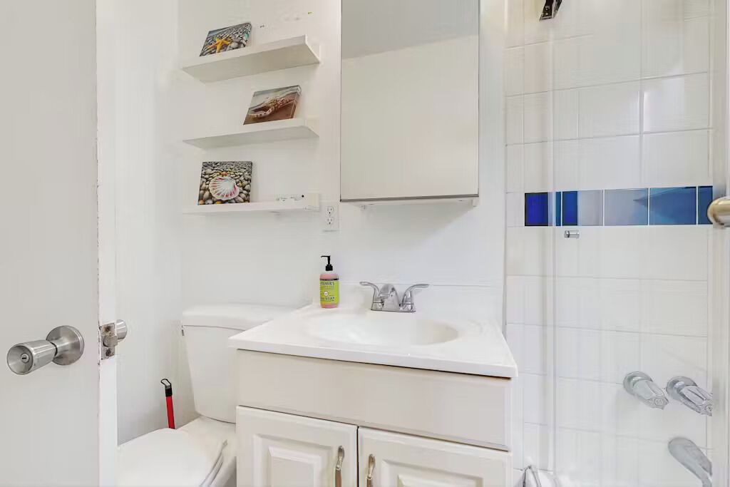 A bathroom with a white sink, a toilet, and a shower area. There are shelves above the sink with decorative items.