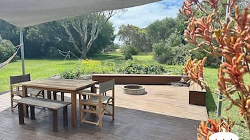 Une terrasse extérieure avec une table et des chaises en bois, un foyer et une vue sur un jardin luxuriant.