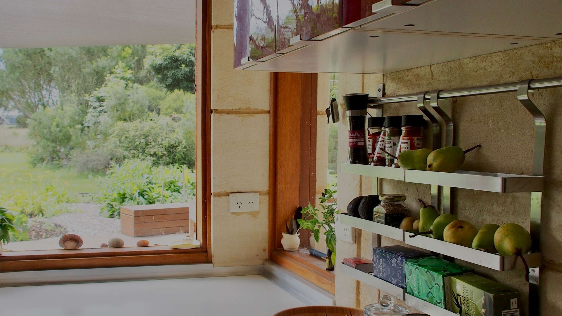 Une cuisine avec une fenêtre donnant sur un espace verdoyant, une étagère avec des bocaux et une plante, et un meuble en bois.