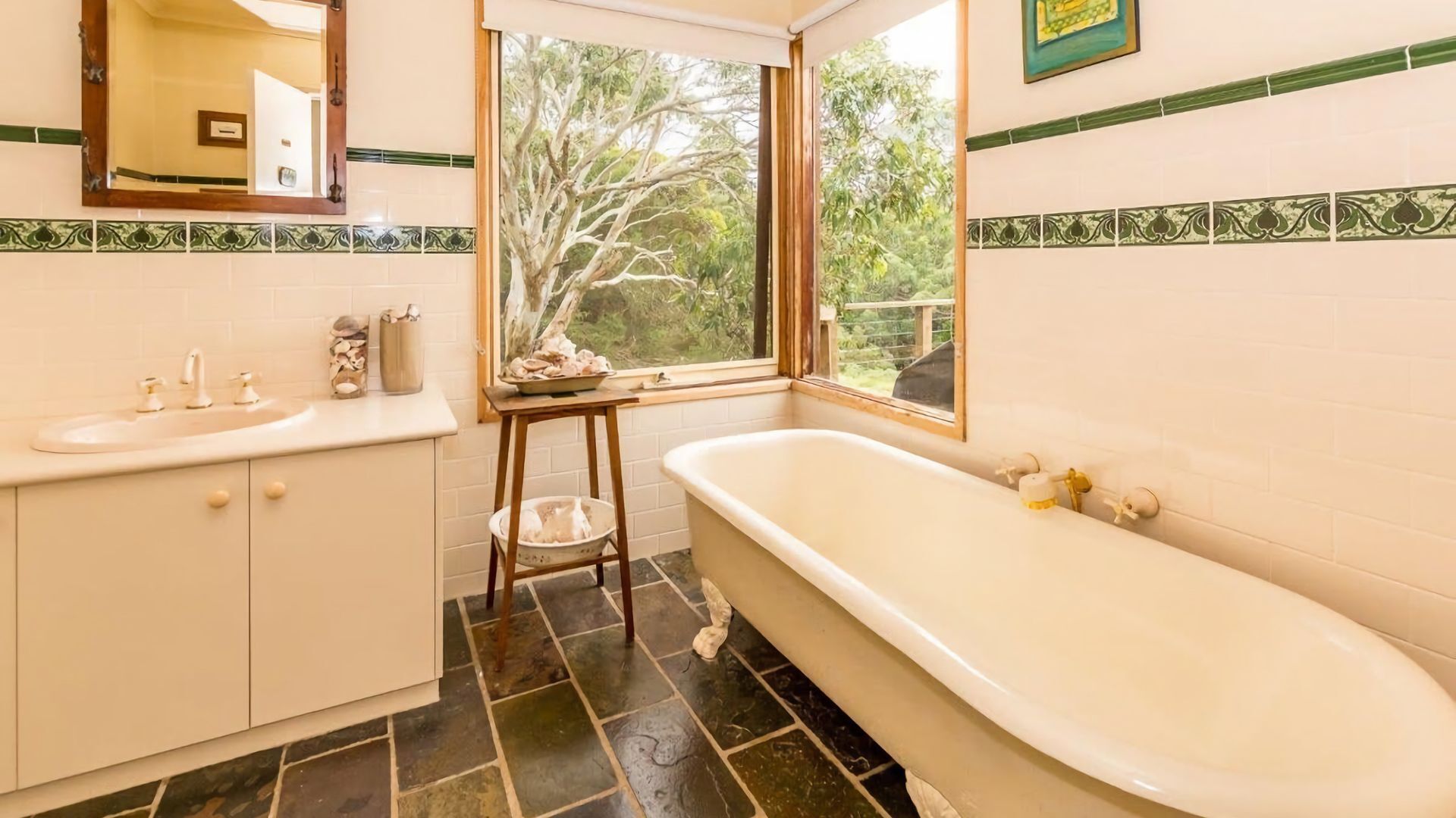 Une salle de bain avec une baignoire, un lavabo, un miroir et une fenêtre donnant sur les arbres.