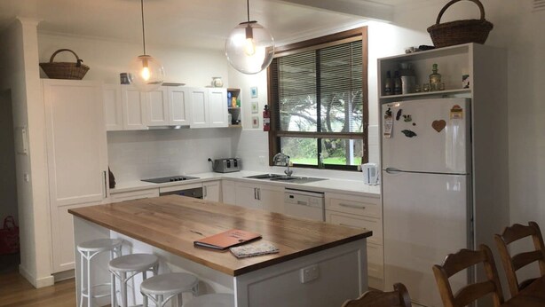 Une cuisine avec des meubles blancs, un îlot central en bois doté de tabourets, une fenêtre avec des stores et un réfrigérateur orné d’aimants.