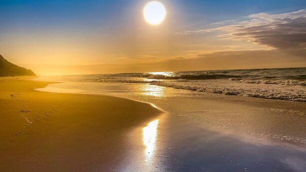 A beach with a clear sky and the sun setting or rising over the ocean.