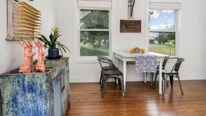 A dining area with a white table, two chairs, a rustic blue cabinet, and a view of the outdoors through two windows. - Smith House | 2/1, Kitchenette, Gas Firepit (Fredericksburg)