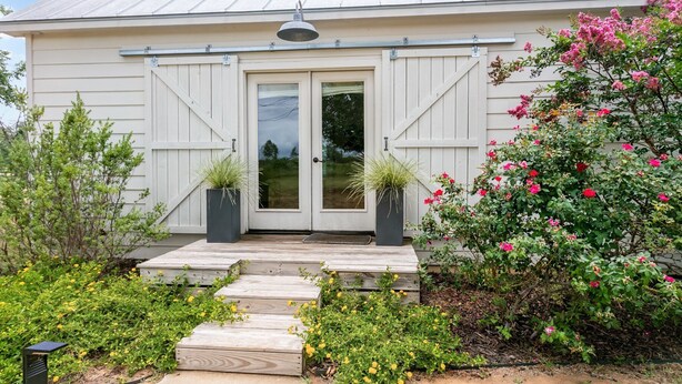 A house with a front door, two potted plants, a wooden porch, and a garden with various plants and flowers. - Smith House | 2/1, Kitchenette, Gas Firepit (Fredericksburg)