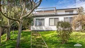 Une maison moderne à deux étages, avec une façade blanche, une terrasse en bois et un petit jardin.