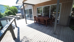 Une terrasse en bois avec une table et des chaises, une porte vitrée donnant sur un espace intérieur et une rambarde en métal.