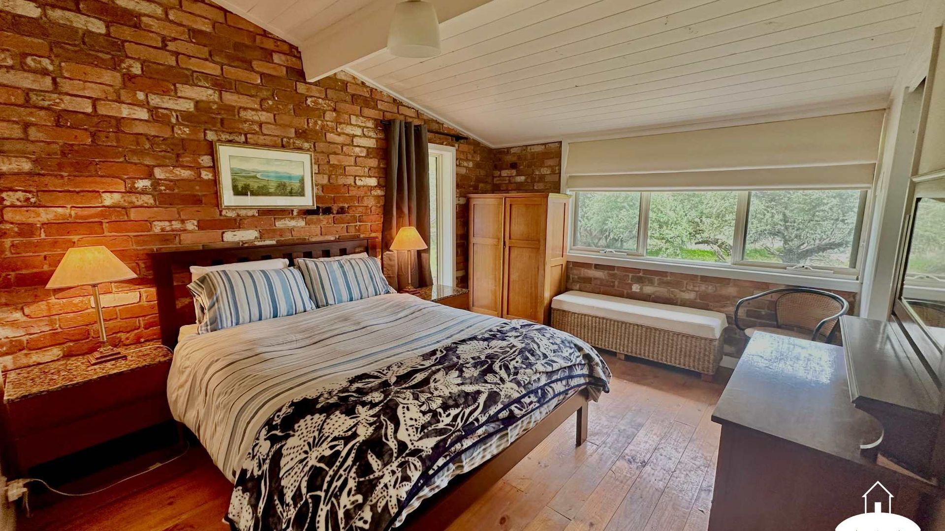 Une chambre avec un mur en briques, un lit avec une literie à rayures, un banc, un bureau et une fenêtre donnant sur des arbres.