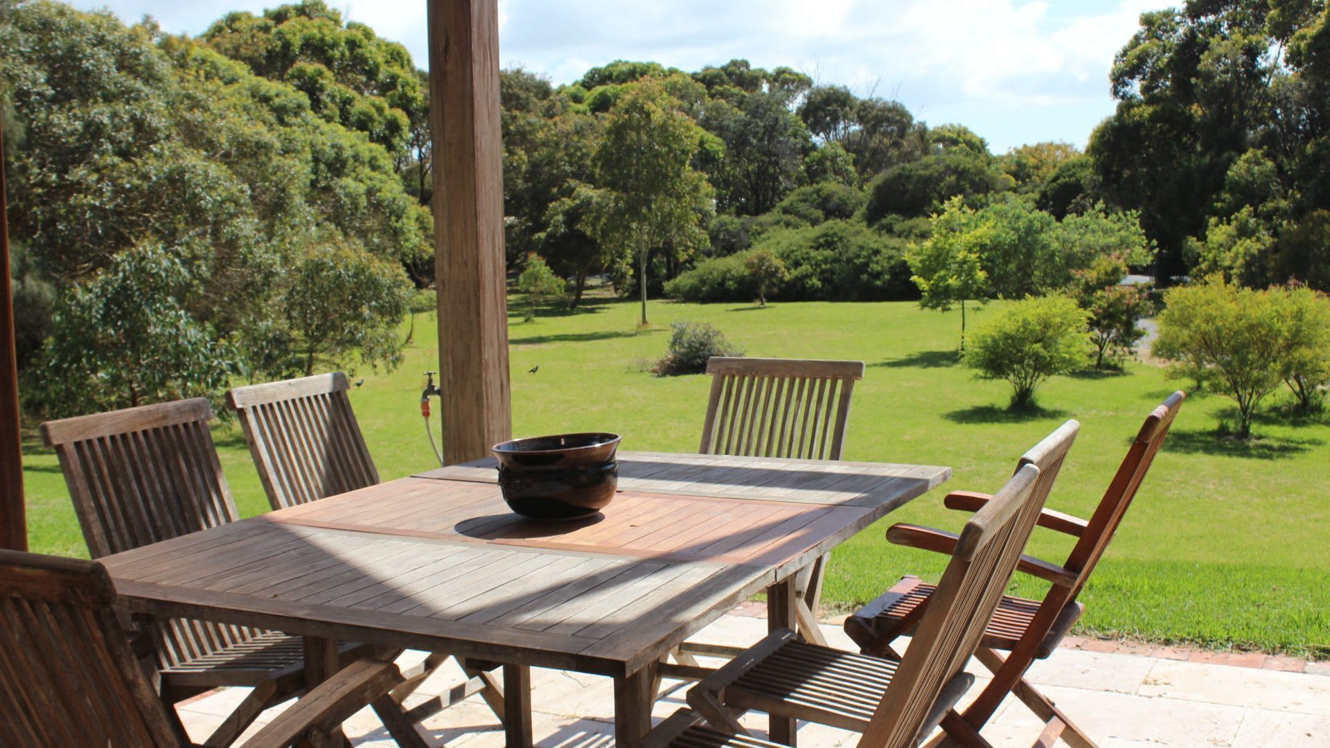 A wooden table with chairs in a garden setting, surrounded by trees and a grassy lawn.