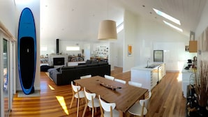 A modern living room with a wooden dining table, white chairs, a black sofa, and a blue surfboard leaning against the wall.