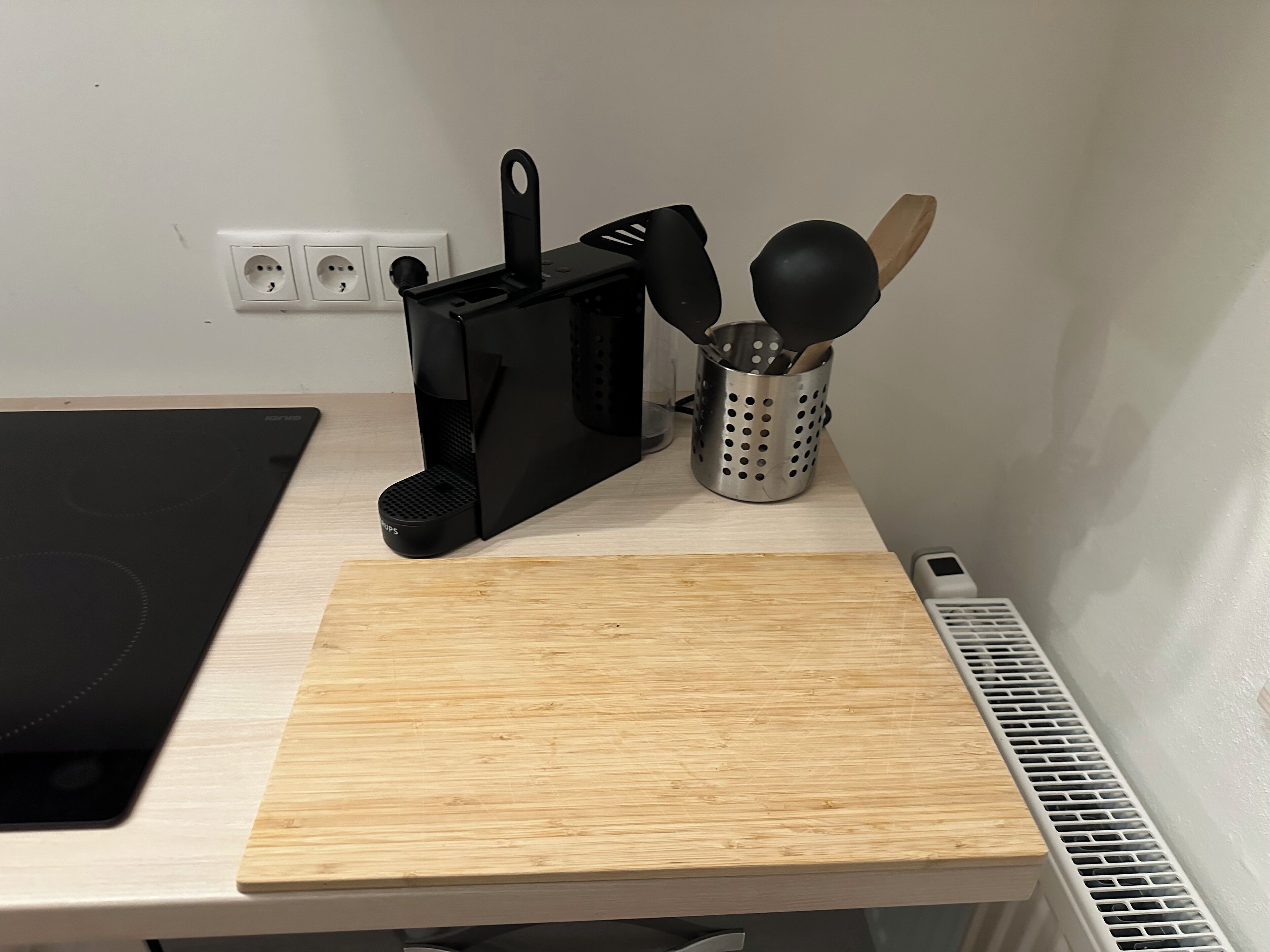 A kitchen counter with a black coffee maker, a wooden cutting board, a stainless steel utensil holder, and a vent.