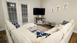Una sala de estar con un sofá seccional beige grande, un televisor de pantalla plana montado en un mueble blanco y una mesita de noche pequeña.