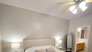 Una cama bien hecha con cabecera capitonada, dos lámparas de noche, un ventilador de techo y un baño visible a través de una puerta abierta.