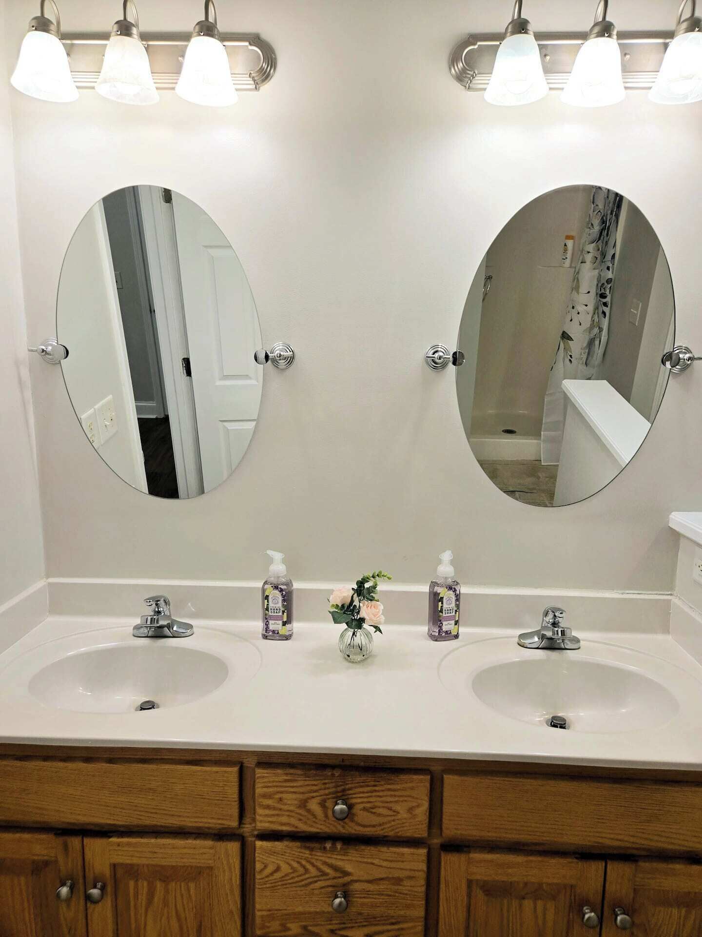 Une salle de bain avec deux lavabos, un miroir et une douche. On trouve deux flacons de gel hydroalcoolique sur le comptoir.