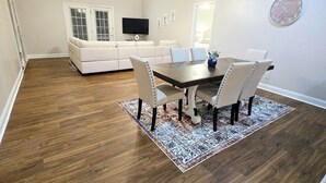 Un comedor con piso de madera, una mesa de comedor con sillas, un sofá, un televisor y un reloj en la pared.