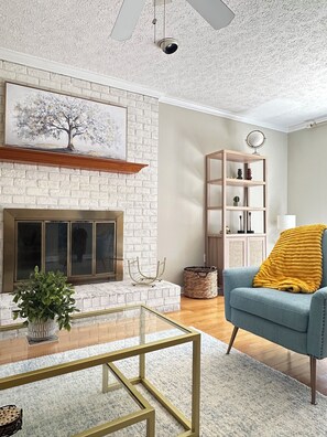 A living room with a white brick fireplace, a glass coffee table, a blue armchair with a yellow throw, and a wooden bookshelf. - Coastal Comfort on Wilmington Island – Minutes to Beach & Downtown (Savannah)