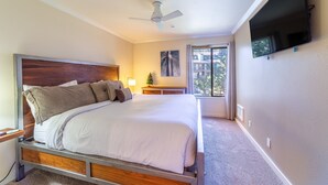 Un dormitorio con una cama grande, un ventilador de techo, un televisor montado en la pared y una ventana con cortinas.