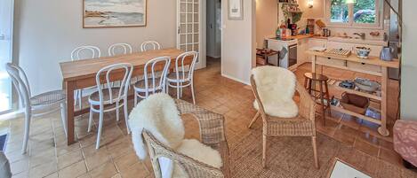 A dining area with a wooden table and chairs, a kitchen with a window, and a living area with a sofa and a white furry chair.