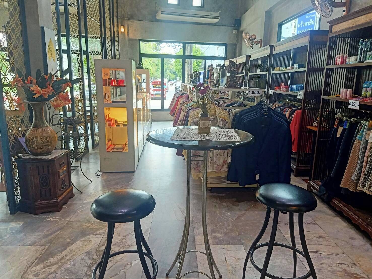 A clothing store with a central round table and two stools, racks of clothes, and a display of vases.
