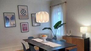 A dining area with a dark wooden table, blue chairs, a white vase with fruits, and framed artwork on the wall.