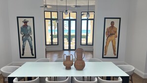 Ein Essbereich mit einem weißen Tisch, sechs Stühlen und zwei gerahmten Kunstwerken, die Personen in westlicher Kleidung zeigen.