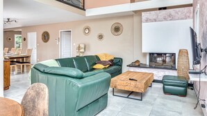 Un vaste salon avec un canapé en cuir vert, une table basse en bois et un pouf vert. On y trouve une cheminée, un coin repas à l’arrière-plan et un grand téléviseur fixé au mur.