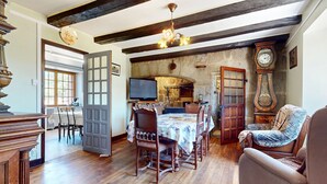 A traditional room with exposed wooden beams, a stone wall, a dining table with chairs, a television, and a large clock.