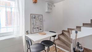 Una habitación pequeña y moderna con un escritorio blanco, dos sillas negras, una obra de arte abstracta enmarcada, un armario de madera y una escalera.