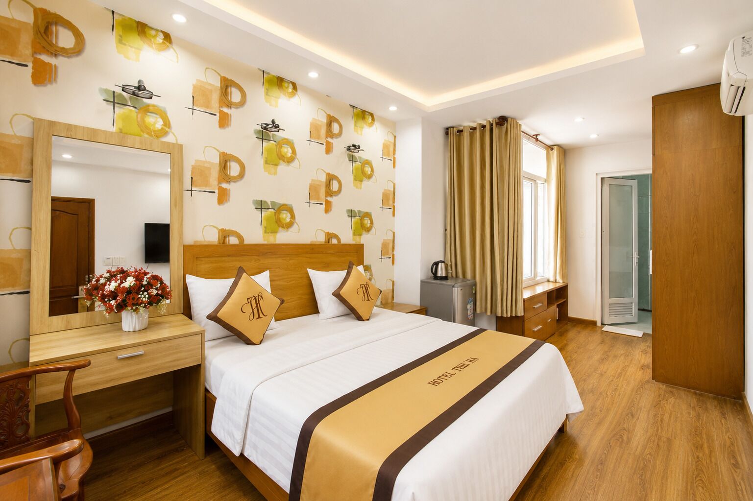 A hotel room with a wooden bed, a desk with a mirror, a small table with a vase of flowers, and a wall with abstract patterns.