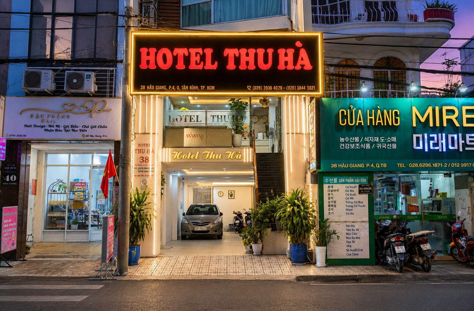 A hotel entrance with a neon sign, a car parked in front, and several potted plants.