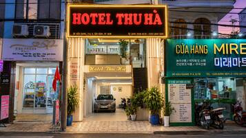A hotel entrance with a neon sign, a car parked in front, and several potted plants.
