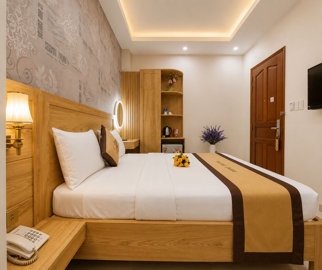A hotel room with a bed, bedside lamp, bedside telephone, a wooden wardrobe, a small shelf with toiletries, a potted plant, and a television mounted on the wall.