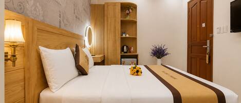 Habitación de hotel con cama, lámpara de noche, teléfono junto a la cama, armario de madera, estante pequeño con artículos de aseo, planta en maceta y televisor montado en la pared.