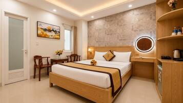 A modern hotel room with a wooden bed, a bedside table with a lamp, a round mirror, and a small seating area with a chair and a table.