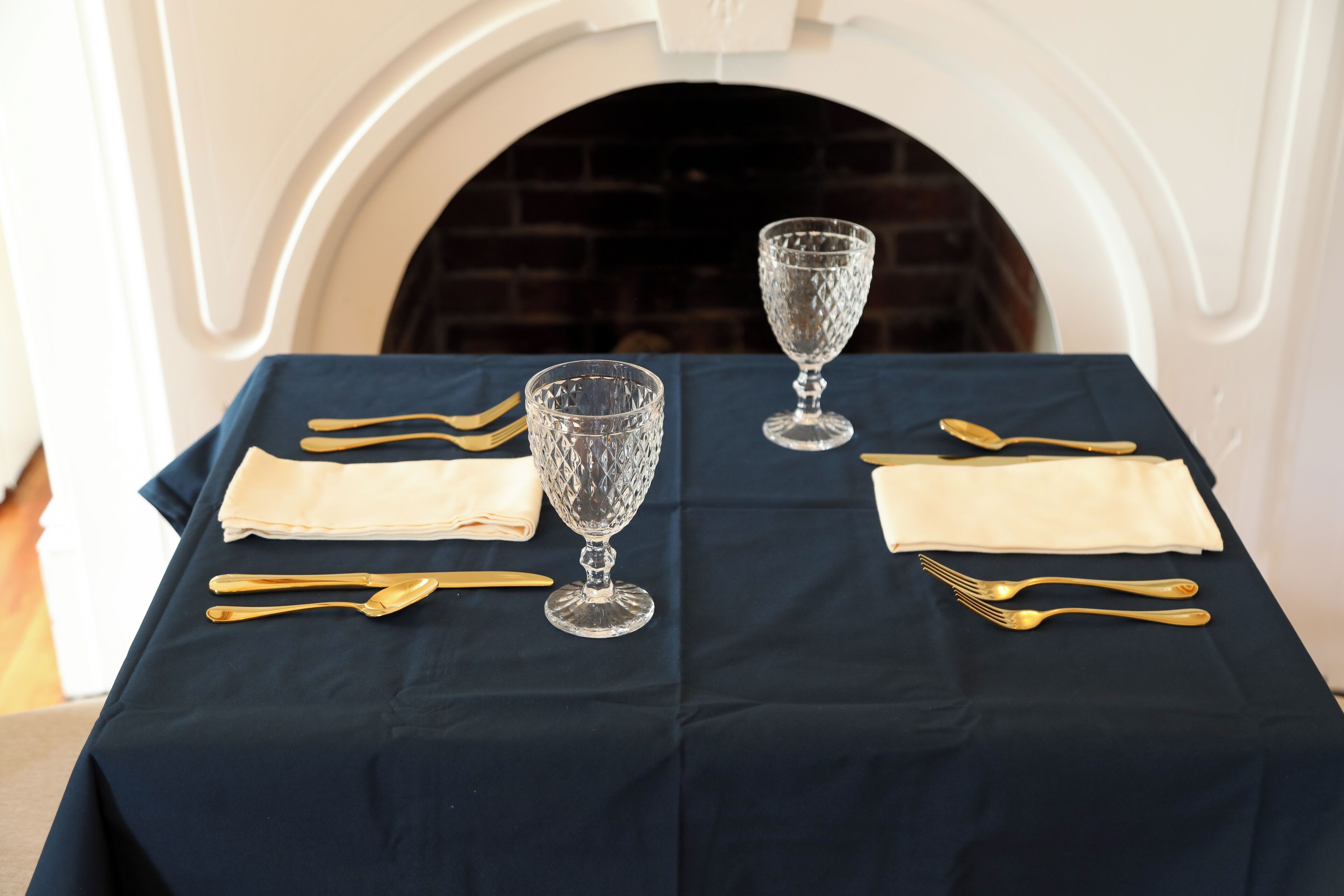 A table set with gold cutlery and crystal glassware, positioned in front of a fireplace.