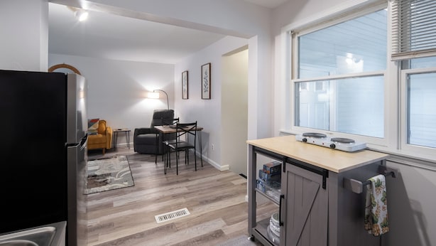 Een compacte keuken met een koelkast, een klein eethoekje met stoelen, en een raam met jaloezieën.